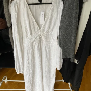 Vit klänning från Gina Tricot - Säljer en elegant vit klänning från Gina Tricot med v-ringning och långa ärmar. Klänningen har ett subtilt broderat mönster och är tillverkad i ett lätt och luftigt material. Perfekt för sommarens alla tillfällen. 🌸