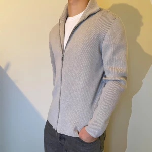 Grå ribbad kofta med dragkedja - Säljer en stilren grå ribbad kofta med dragkedja. Perfekt för lager-på-lager och har en bekväm passform. Koftan har långa ärmar och en hög krage som ger extra värme.