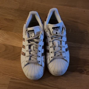 Adidas Superstar med leopardmönster - Snygga vita Adidas Superstar sneakers med leopardmönstrade detaljer på sidorna och hälen. Klassisk snörning och den ikoniska tåhättan i gummi. Perfekta för att ge din stil en trendig touch.