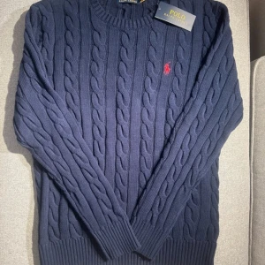 Mörkblå kabelstickad tröja från Polo Ralph Lauren - Säljer en stilren mörkblå kabelstickad tröja från Polo Ralph Lauren. Tröjan har en klassisk design med rund hals och det ikoniska röda logotypbroderiet på bröstet. Perfekt för kyligare dagar!