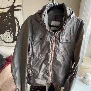 Grön vindjacka från Moncler i storlek S - Snygg vindjacka från Moncler med huva och dragkedja i rött Jackan har två bröstfickor med knappar och Moncler-logga på ärmen. Pris kan diskuteras!