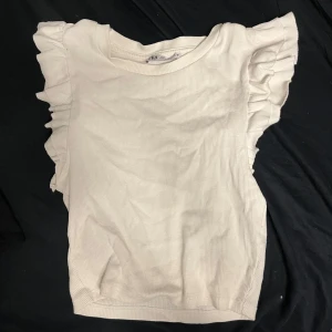 Vit ribbad topp med volangärmar - Säljer en söt vit ribbad topp från H&M med volangdetaljer på ärmarna. Perfekt för en stilren och chic look. Toppen har en rund halsringning och är i ett mjukt material.