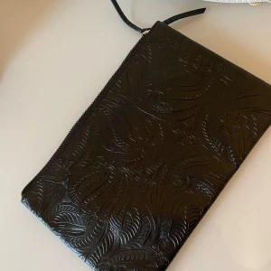 Svart clutchväska från Pieces - Snygg elegant svart clutchväska från Pieces med ett subtilt mönster och dragkedja. Perfekt för att bära med sig det viktigaste. Bred: 31cm och höjd : 21cm . Liten på kanten har tappat  tyget.syns på sista bild 