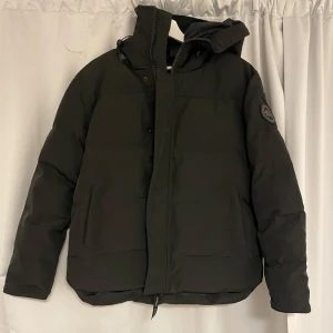 Svart dunjacka från Canada Goose - Säljer en svart macmillan med svart label från Canada Goose med huva och dragkedja. Jackan har en klassisk design med långärmade ärmar och en logotyp på ärmen. Perfekt för kalla dagar. Ps den har inte använts helt ny med Canada Goose väskan.