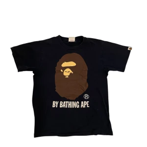 marinblå A Bathing Ape t-shirt - Snygg marinblå t-shirt från A Bathing Ape med det ikoniska aptrycket i brunt och texten 'BY BATHING APE' på framsidan. Perfekt för streetwear-entusiaster.