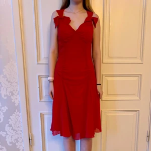 Klänning med rosett - Jättefin röd klänning i skönt material med två fina rosetter. Storlek  38 Modellen på bilden är 173 cm och har storlek 38 i de flesta andra kläder. Aldrig använd. Vi har lagt ner vår klänningsbutik och säljer ut. Ny pris är 2000-3000kr