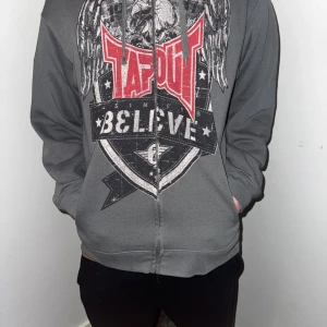 Grå hoodie från Tapout - Säljer en grå hoodie från Tapout med tryck i rött och svart. Hoodien har en dragkedja framtill och en stor grafisk design med texten 'Believe'. Perfekt för en avslappnad stil.