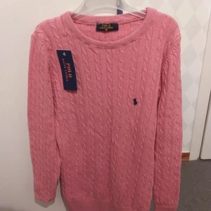 Rosa kabelstickad tröja från Polo Ralph Lauren - Säljer en snygg rosa kabelstickad tröja från Polo Ralph Lauren. Tröjan har en klassisk design med rund hals och det ikoniska logotypbroderiet på bröstet. Perfekt för en stilren look!     Min sister fick den i julklapp av sin vän men den var för liten för henne, den är helt oanvänd och etiketten är fortfarande på. Storleken är dam M men det passar bättre om man har dam S.