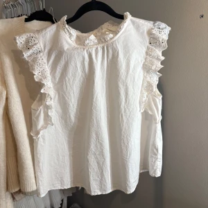 Vit blus med spetsdetaljer - Charmig vit blus med vackra spetsdetaljer på ärmarna och runt halsen. Blusen har en luftig passform och knäppning i nacken. 