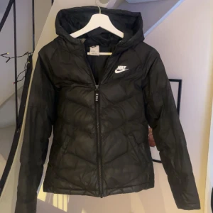 Svart pufferjacka från Nike - Säljer en svart pufferjacka från Nike med huva och dragkedja. Jackan har en stilren design med Nike-logga på bröstet. Perfekt för kyligare dagar.