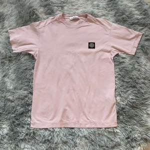 Rosa t-shirt från Stone Island - Snygg rosa t-shirt från Stone Island med korta ärmar och en klassisk rund halsringning. T-shirten har en diskret logotyp på bröstet och är tillverkad i mjukt bomullsmaterial.