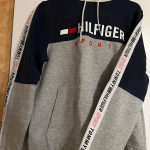 Grå och marinblå hoodie från Tommy Hilfiger - Säljer en hoodie från Tommy Hilfiger i grått och marinblått med logga på bröstet och längs ärmarna. Storlek XS.