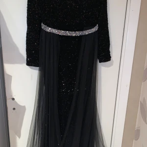 Svart glittrig långklänning med paljetter - Elegant svart långklänning med glittriga paljetter och ett dekorativt bälte i midjan. Klänningen har långa ärmar och ett skirt lager över kjolen som ger en fin effekt. Perfekt för speciella tillfällen.