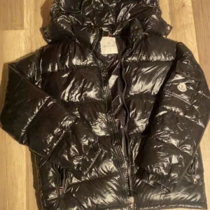 Svart dunjacka från Moncler - Säljer en snygg svart dunjacka från Moncler med glansig finish. Jackan har en praktisk huva och dragkedja framtill. Perfekt för kyliga dagar med stil.