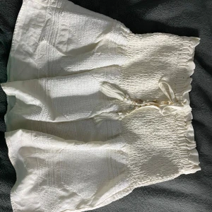 Zara kjol - Charmig vit kjol med elastisk midja och knytband framtill. Kjolen har en smockad design som ger en fin textur och volym. Perfekt för varma dagar. Kjolen var populär bland zaras gamla kollektion