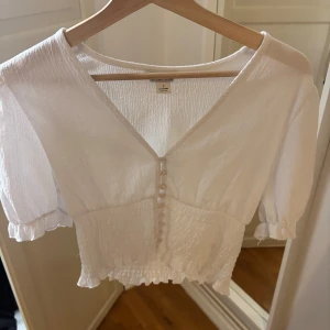 Vit blus från Monki - Säljer en söt vit blus från Monki med korta puffärmar och knappar framtill. Blusen är croppad och har en v-ringad design med elastiska detaljer vid ärmslut och nederkant. Använd fåtal gånger!
