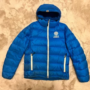 Blå dunjacka från Franklin Marshall - Säljer en snygg blå dunjacka från Franklin Marshall med huva och dragkedja. Jackan har vita detaljer och två praktiska fickor med dragkedja framtill. Perfekt för kyliga dagar!