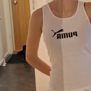 Vit linnetopp från Puma - Säljer en vit linnetopp från Puma med svart logga framtill. Perfekt för träning eller en avslappnad dag. Toppen har en klassisk rund halsringning och ärmlös design. Är I storlek XS