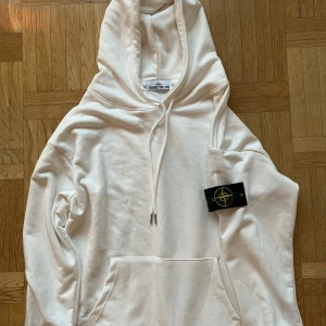 Vit hoodie Stone Island - Säljer en stilren vit hoodie från Stone Island med klassisk logga på ärmen. Hoodien har en rymlig ficka framtill och justerbar dragsko i huvan. Perfekt för en avslappnad look.