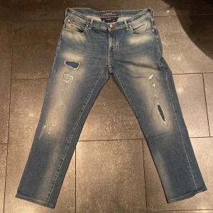 Blå jeans från Jacob Cohen - ÄKTA Snygga blå jeans från Jacob Cohen med slitna detaljer och broderad logga. De har en klassisk femficksdesign och knappar i midjan. Perfekta för en avslappnad stil med en touch av lyx. Italiensk storlek 30 