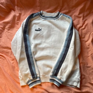 Puma Sweatshirt  - Säljer en vit tröja från Puma med blå ränder längs ärmarna och ribbade muddar. Tröjan har en klassisk design med rund hals och Puma-logga på bröstet. Perfekt för en avslappnad stil. En mindre fläck som syns på sista bilden. 