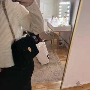 Svart axelväska med kedja - Snygg svart axelväska i sammet med guldfärgad kedja och rund låsdetalj. Perfekt för att ge en elegant touch till din outfit. Väskan har ett rymligt fack och en stilren design.