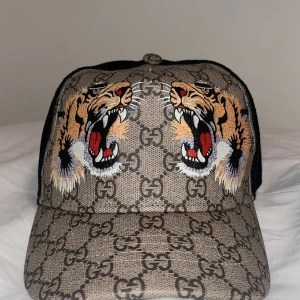 Gucci keps med tigerbroderi Size M - Snygg keps från Gucci med ikoniskt GG-mönster och broderade tigrar framtill. Kepsen har ett justerbart band baktill i grönt och rött. Perfekt för att ge din outfit en lyxig touch.