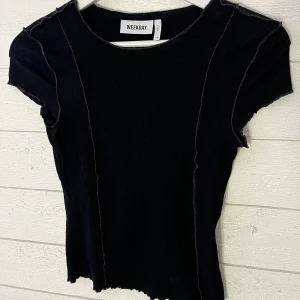 Svart ribbad topp från Weekday - Snygg svart ribbad topp från Weekday med korta ärmar och råa kanter. Perfekt för en stilren look. Toppen har en tight passform och är tillverkad i ett mjukt material.