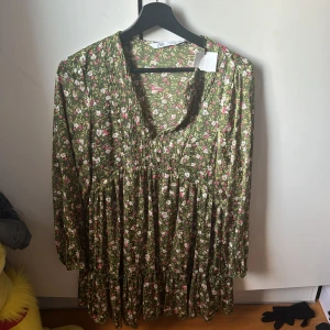 Blommig klänning från Zara - Charmig grön klänning från Zara med ett blommigt mönster i rosa och vitt. Klänningen har en v-ringad design och långa ärmar, perfekt för en avslappnad stil. Tillverkad i ett lätt och bekvämt material. Prislappen är kvar!