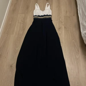 Vit och marinblå långklänning - Elegant långklänning med vit överdel och marinblå kjol. Klänningen har spetsdetaljer i midjan och en djup v-ringning. Perfekt för formella tillfällen.