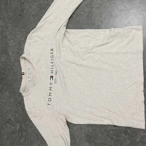 Vit långärmad tröja från Tommy Hilfiger - Snygg vit långärmad tröja från Tommy Hilfiger med tryck på framsidan. Perfekt för en stilren och avslappnad look. Tröjan har en klassisk rund halsringning.