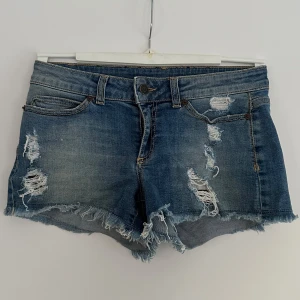 Lågmidjade jeansshorts med slitningar - Snygga blå jeansshorts med coola slitningar och fransiga kanter från Hunkedory. Midjemått tvärs över: 35 cm. 