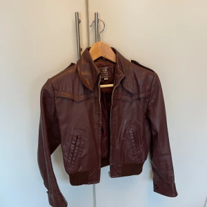 Rödbrun/vinröd skinnjacka från H&M - Snygg skinnjacka från H&M i äkta läder. Jackan har en klassisk krage och stängs med dragkedja. Den har långa ärmar med knappar vid ärmsluten och ribbade muddar i nederkant. Perfekt för en cool och stilren look. Storlek 36 men passar nog XS/32/34. Ser brun ut på bilden men är mer vinröd på riktigt. Pris kan diskuteras!