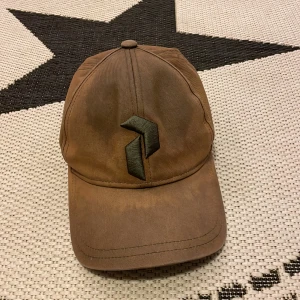 Brun Peak Performance keps med justerbart spänne - Snygg brun keps med ett svart broderat märke framtill. Kepsen har ett justerbart spänne baktill för perfekt passform. Perfekt för en avslappnad stil.