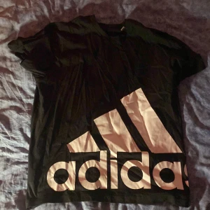 Svart t-shirt från Adidas - Snygg svart t-shirt från Adidas med stort logotryck i vitt på framsidan. Perfekt för en sportig look eller avslappnad stil. Kortärmad och bekväm passform.