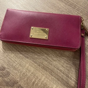 Vinröd/lila plånbok från Michael Kors - Snygg vinröd/lila plånbok från Michael Kors med guldfärgad dragkedja och logotypplatta. Plånboken har flera fack för kort och sedlar, samt en avtagbar handledsrem. Perfekt för att hålla ordning på dina värdesaker med stil. Köp för 2000 inga defekter🩷🩷