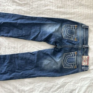 Blå jeans från True Religion - Snygga blå jeans från True Religion med kontrasterande vit söm och klassisk femficksdesign. Jeansen har en låg midja och en straight passform, perfekt för en avslappnad stil. Märkeslogga på bakfickan och knappar i metall för en cool detalj.