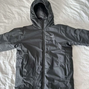 Svart dunjacka från Arc'teryx - Säljer en svart dunjacka från Arc'teryx med huva och dragkedja. Perfekt för kyliga dagar med sin stilrena design och funktionella passform.
