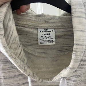 Gråmelerad hoodie från Champion - Säljer en gråmelerad hoodie från Champion med justerbar huva och känguruficka. Perfekt för en avslappnad stil. Den har långa ärmar och ett tryck på bröstet.