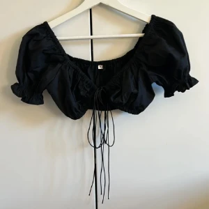 Svart kortärmad topp med knytning - Trendig svart topp med korta puffärmar och knytning framtill. Aldrig använd, i nyskick. Tryck gärna på ”köp nu”.