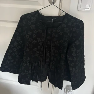 Svart blommig blus från Vero Moda - Snygg svart blus från Vero Moda med blommigt mönster och knytband framtill. Blusen har en normal passform och är perfekt för höst och vår. Den har långa ärmar och en unik design som sticker ut.