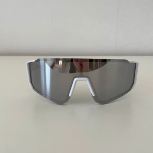 Sportiga solglasögon med spegelglas - Snygga sportiga solglasögon med spegelglas och vit ram. Perfekta för utomhusaktiviteter med en modern och stilren design. Glasögonen har en omslutande passform som ger bra skydd mot solen.