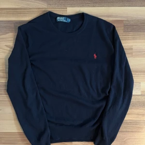 Mörkblå tröja från Polo Ralph Lauren - Snygg mörkblå tröja från Polo Ralph Lauren med röd broderad logga på bröstet. Tröjan har en klassisk rund hals och långa ärmar, perfekt för en stilren look. Lite sprättning syns på sista bilden.