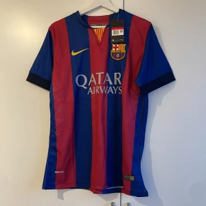 FC Barcelona fotbollströja med Messi 10 - Snygg FC Barcelona tröja från Nike med Messi 10 på ryggen. Tröjan är randig i rött och blått med klubbens emblem och Qatar Airways logga på framsidan. Kortärmad design med Dri-FIT teknologi för optimal komfort. Barca hemma tröja 2014/2015