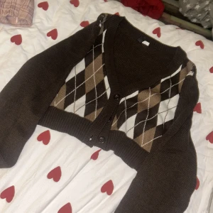 Brun kofta med argylemönster från H&M - Säljer en brun kofta från H&M med klassiskt argylemönster i brunt, beige och vitt. Koftan har knappar framtill och är perfekt för en stilren look.  den är croppas också