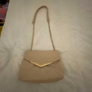 Beige axelväska med kedja - Snygg beige axelväska med guldfärgad kedja och dekorativt V-format spänne. Väskan har ett quiltat mönster och en praktisk dragkedja inuti för extra säkerhet. Perfekt för att bära dina nödvändigheter med stil.