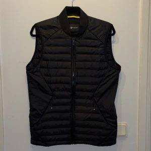 Svart dunväst från Sail Racing - Snygg svart dunväst från Sail Racing med dragkedja framtill. Västen har en stilren design med quiltade paneler. Den är lätt och smidig att bära, vilket gör den till ett mångsidigt plagg i garderoben. Perfekt för kommande vår. Använt i en säsong endast, som ny, i princip defektfri (endast på dragkedjan, se bild 3)