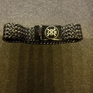 Svart armband med metallspänne - Snyggt svart armband med ett flätat mönster och ett metallspänne med logotyp. Perfekt accessoar för en stilren look.