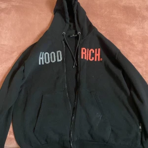 Svart hoodie från Hoodrich - Säljer en svart hoodie från Hoodrich med texten 'HOOD RICH' i grått och rött på bröstet. Den har en dragkedja framtill och en justerbar huva med snören. Perfekt för en avslappnad stil. Storleken är XL men passar även L. Pris kan diskuteras köptes för 1200kr ordinarie pris.