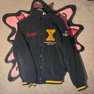 Mörkblå varsity jacka  - Snygg jacka med broderade detaljer. Jackan har en stor gul bokstav 'X' på bröstet och texten 'New York' i rött. Den har knappar framtill och ribbade muddar i svart och gult.